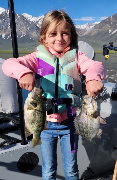 a child standing on a boat holding two Sacramento perch with snow capped mountains in the background