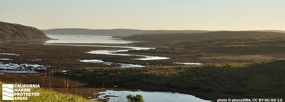 rounded brown cliffs roll into a windy snake-like estuary, the blue sky is contrasted with the golden glow of sunset along the horizon, this reflection makes the estuary glow blue contrasting the brown land around it