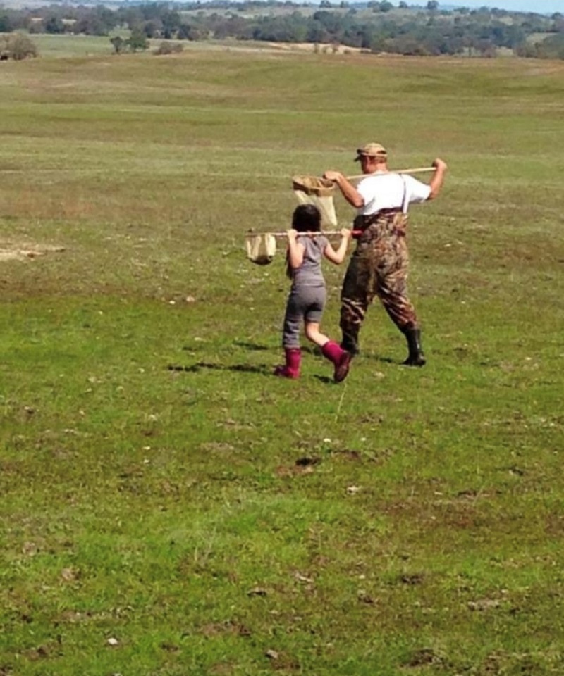 An adult and a child walking across a wetland wearing boots and holding dipnets on their shoulders.