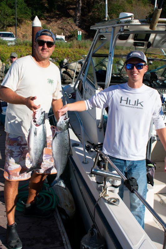 Two men, one on a boat and one on a dock, each hold a salmon