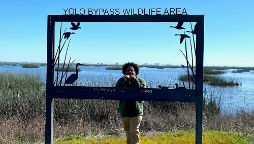 CDFW Seasonal Aid Darian Marico Clark-Stinson stands behind the Yolo Bypass Wildlife Area metal sign.