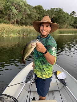 Farhat Bajjaliya holding up a largemouth bass while fishing, lake in the background.