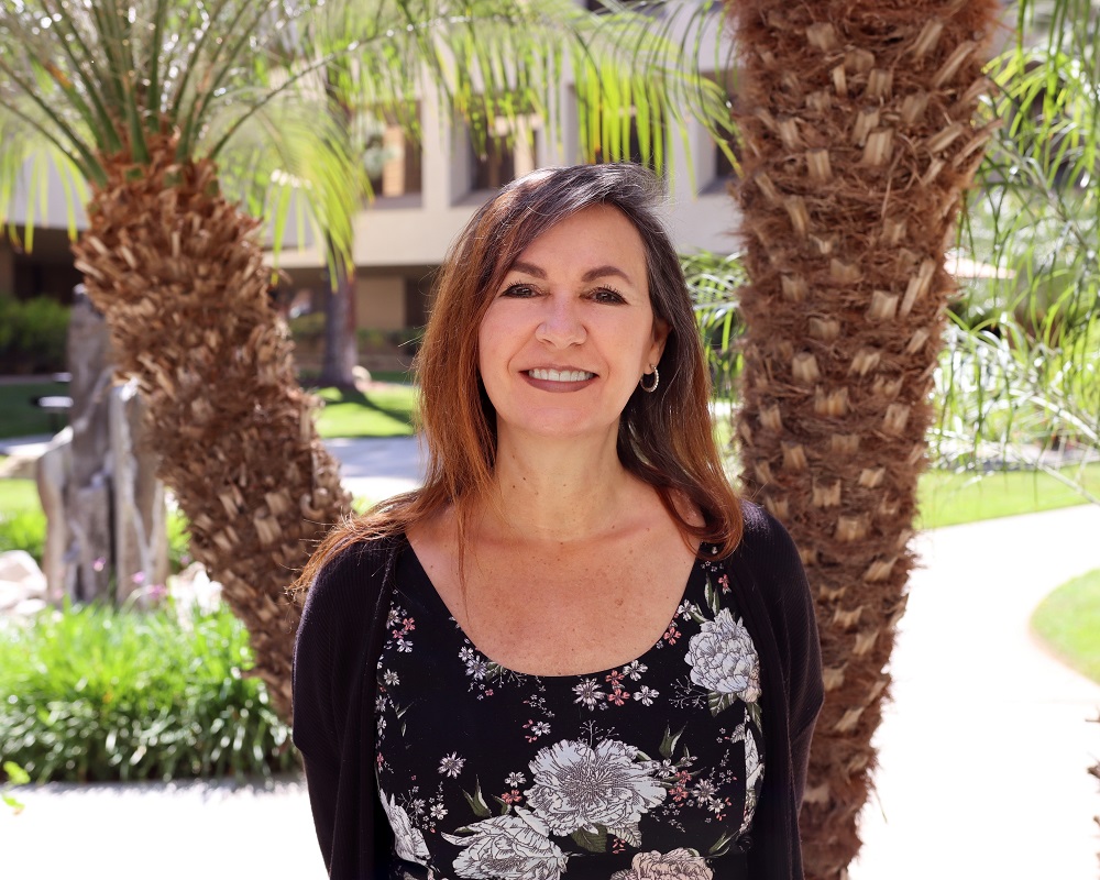 Outdoor headshot of Karen Wold, wearing a black patterned blouse and smiling, standing in front of a double palm tree
