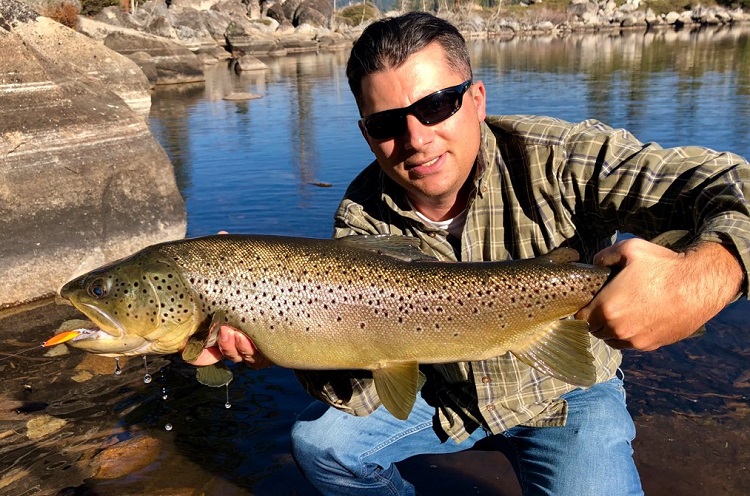 An angler holds up a large brown trout caught with a lure in a mountain lake.
