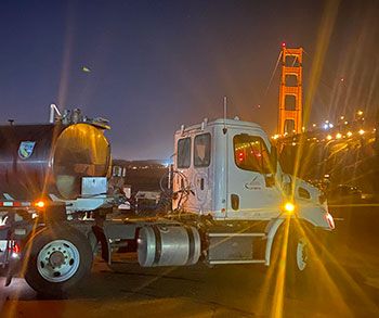 A silver CDFW fish stocking tanker truck is parked in front of the Golden Gate Bridge at night, with lights from the bridge reflecting off of the truck.