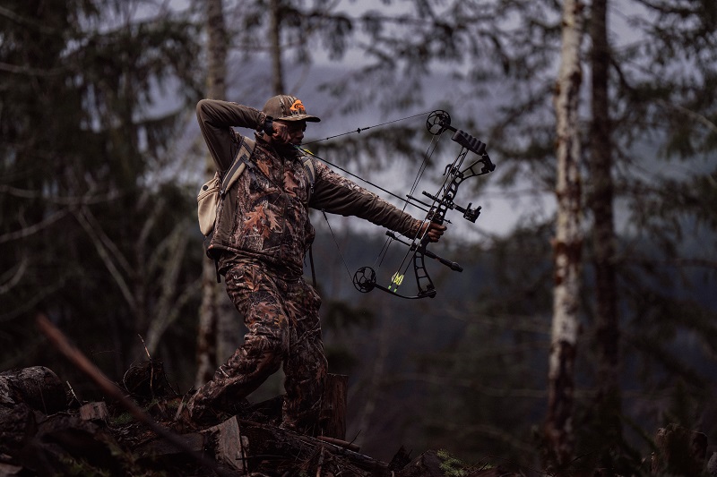 A camouflage-outfitted big game hunter pulls back his bow and takes aim in the forest.