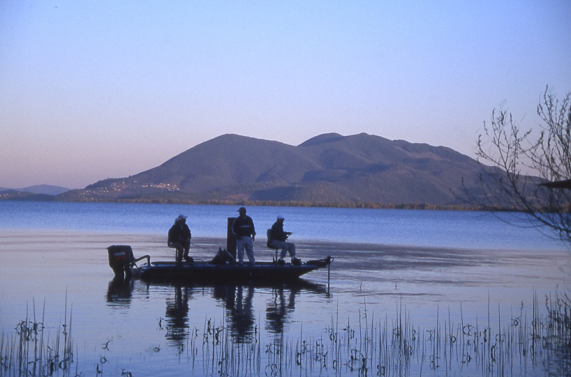 Three men fishing on boat in water. Large mountain in background. Fishing rods and lines in water.