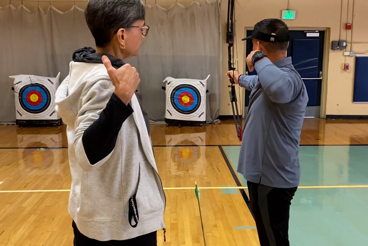 Two people stand in front of an archery target, one aiming a bow.