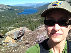 from a mountain pass, a view of an Alpine valley, lake, and more mountains, behind a woman's face