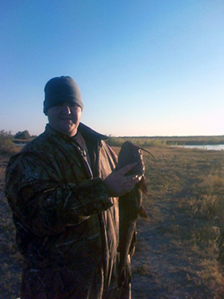 A man stands near a waterway, holding a large catfish under a blue sky