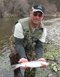 A man wearing camouflage waders hold a silvery fish on a riverbank