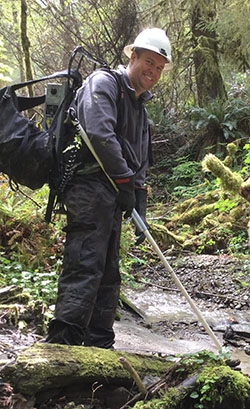 Smiling man in dark grey pants and sweatshirt, black gloves, and white hard hat wearing backpack machine with long pole attached standing in wet, forested area