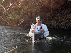 A man wearing waders stands waist-deep in a flowing stream, holding a long, narrow measuring device