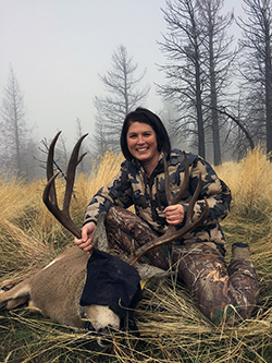 a young woman wearing camouflage kneels on a foggy hillside with an immobilized buck.