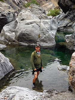 man standing in river pool surrounded by large boulders and hillside