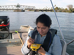 Scientist Shruti Khanna on a boat on the delta taking notes with a bridge in back
