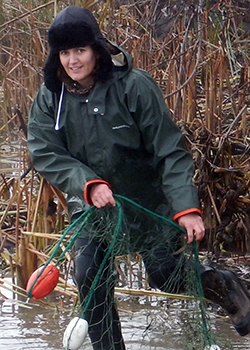 a woman wearing a green, thermal jumpsuit stands in shallow water, pulling a fish net from the marsh
