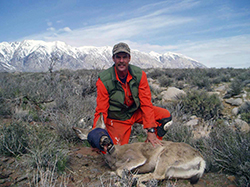 Man in orange jumpsuit kneels in sagebrush with a deer that's hobbled and blindfolded