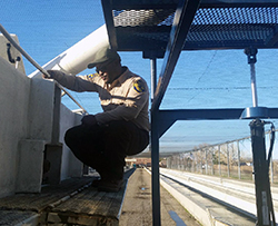 An African American man in a California Fish and Wildlife uniform squats to read something on a low concrete wall at a fish hatchery
