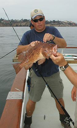 Man standing on boat holding large brown fish while holding fishing pole in crook of arm. Another hand is holding onto the mouth of the fish.