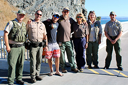 Group of seven people, two women, five men standing on road posing for photograph. Rock face, beach, and body of water in background.