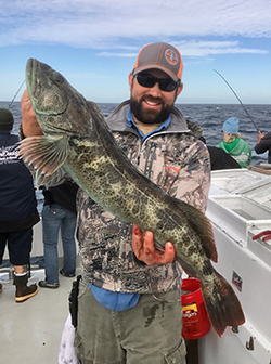 Bearded man wearing camo jacket, green cargo pants, sunglasses, and orange and gray baseball cap holding large lingcod fish on boat in water. People fishing on the boat in the background.