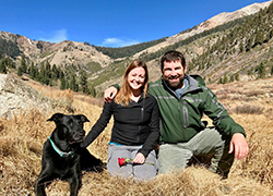 Bearded man in green jacket and green pants kneeling on ground with arm around kneeling woman wearing black jacket and gray pants holding a red rose in one hand and other hand on black dog laying in long dry grass. Mountains and blue sky in background.