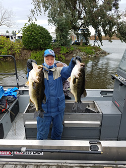 Man wearing blue foul weather gear, blue ball cap, on docked fishing boat holding up two large fish. Water, land, and trees in background.