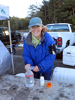 Smiling woman wearing blue hat and blue jacket holding plastic bag, hunched over table surface with glass jar and container. Vehicles and trees in background
