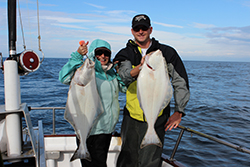 White male wearing grey, black, and yellow jacket and black pants holding up large halibut standing next to white female wearing black pants and blue jacket with hood up holding large halibut. Both are standing on a boat on the water. Partly cloudy sky in background.