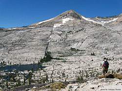 Large sheer rock mountainside with snow at peak and some trees. Man standing on rock appearing very small compared to the mountain.