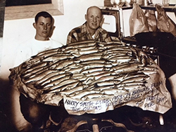 Sepia colored image of young man and older man sitting behind table covered in nearly 100 fish. A sign in front reads Harry Smith, 23 years 1953 74 browns, from the owens river gorge, limit 25 trout per day.