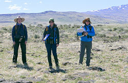 Three scientist standing on dry grass with mountains and blue sky