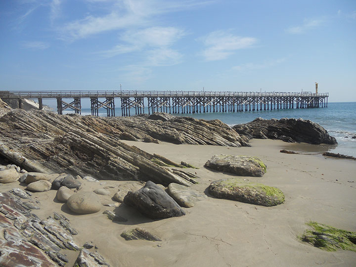 a wooden pier extends along the horizon, round moss covered and angular sandstone rocks extend from the sand meeting the ocean and pier