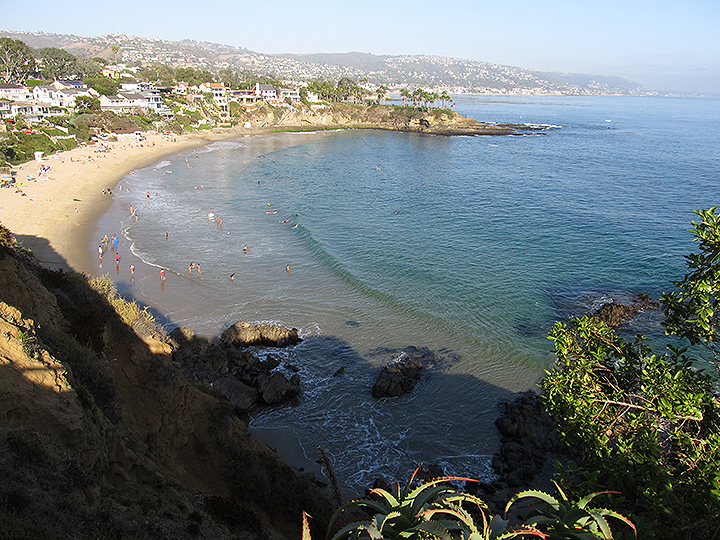 a curved sandy beach meets a gentle ocean, a few dozen beachgoers on the sandy shore, houses stretch up from the beach all the way up a rolling hillside along the horizon