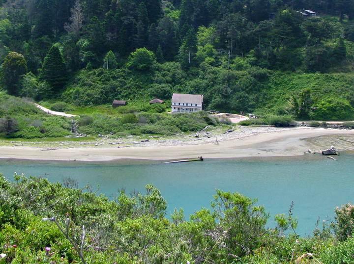 a two-story building on a riverbank below forested hillside