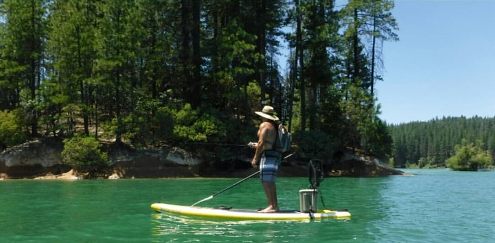 person standing up on paddleboard on water while fishing. Trees in the background.