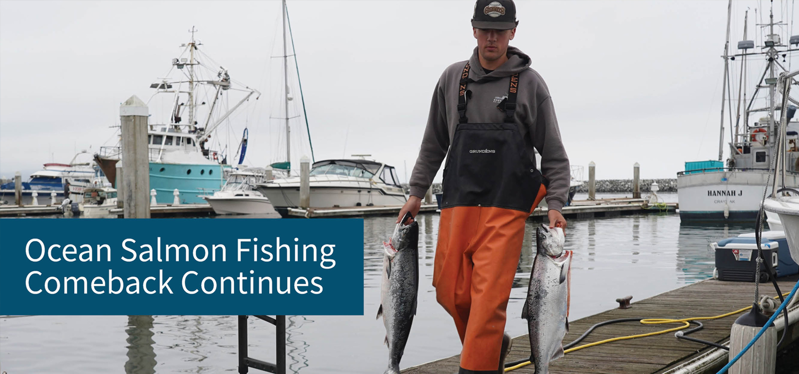 Fisherman holding salmon on a dock with boats in the background