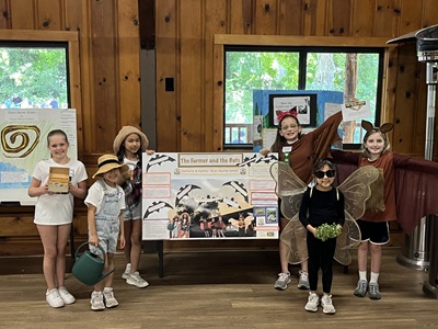 children stand indoors room, wearing costumes and holding craft items. The board features illustrated scenes and text, with windows showing trees in the background
