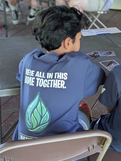 Child sitting at a table during an outdoor activity, wearing a blue shirt with a water droplet graphic and the words “WE’RE ALL IN THIS BIOME TOGETHER