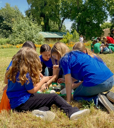 Several children and teens in blue shirts sit together on the grass outdoors, gathered around food or supplies, with trees, flowers, and other people in the background on a sunny day