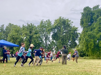 Group of people participating in an outdoor activity on a grassy field, with trees and a blue canopy tent in the background.